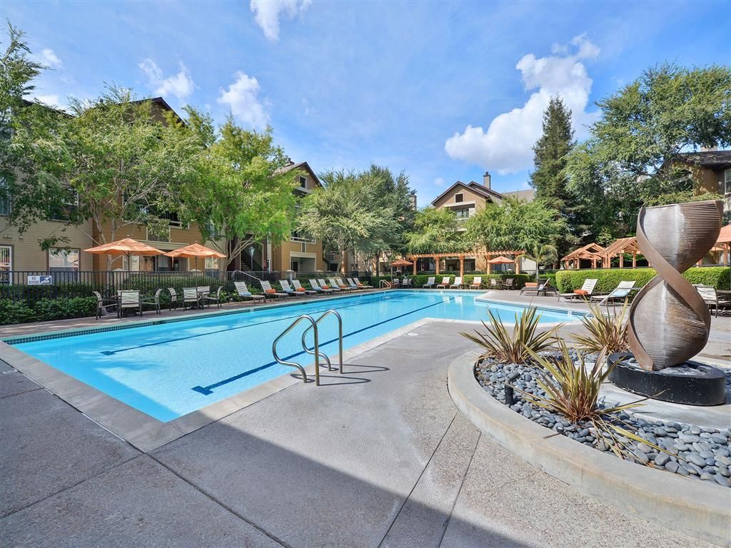 Outdoor community pool with lounge chairs, umbrellas, and landscaping.