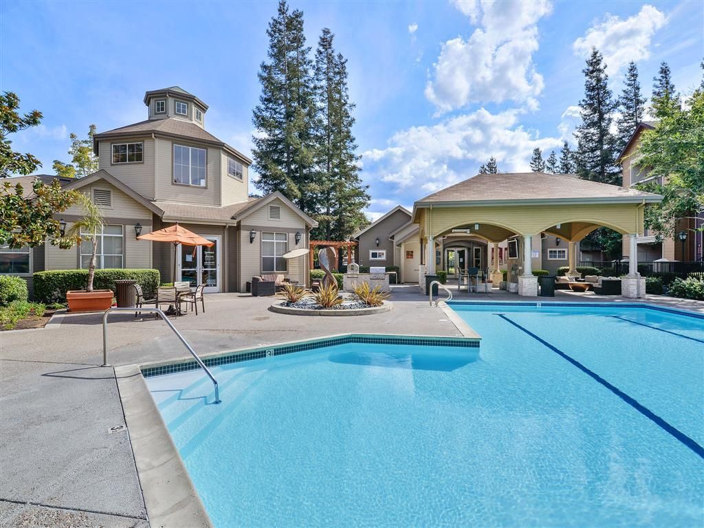 Outdoor apartment community pool with lounge chairs, umbrellas, and a clubhouse in a landscaped courtyard.