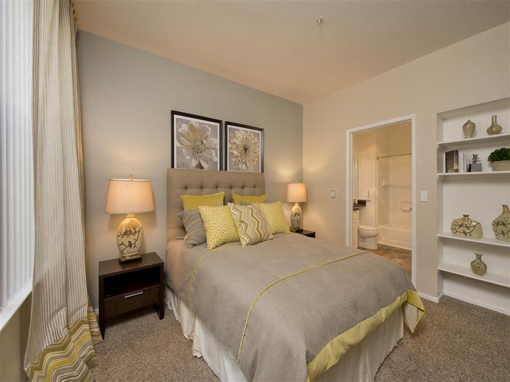Bedroom in an apartment with a neatly made bed, two bedside lamps, and an open doorway to the bathroom.