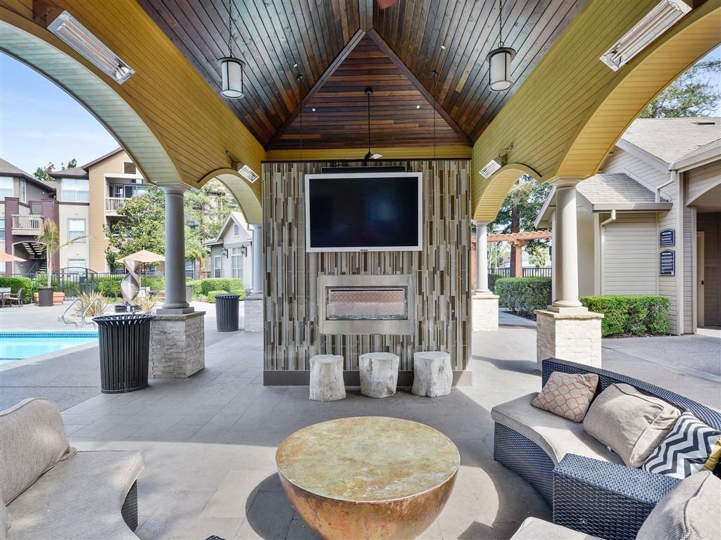 Outdoor community lounge under a wooden pavilion with a TV, fireplace, and seating near the pool.