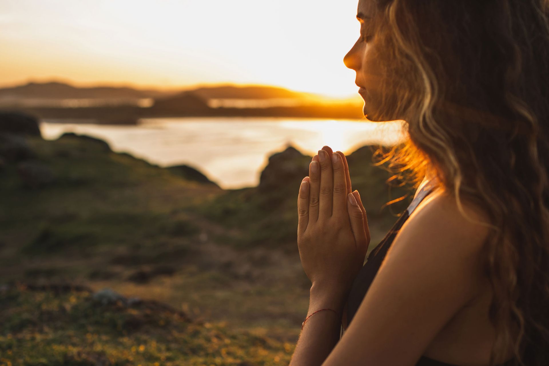 A woman is sitting on a hill with her hands folded in prayer.