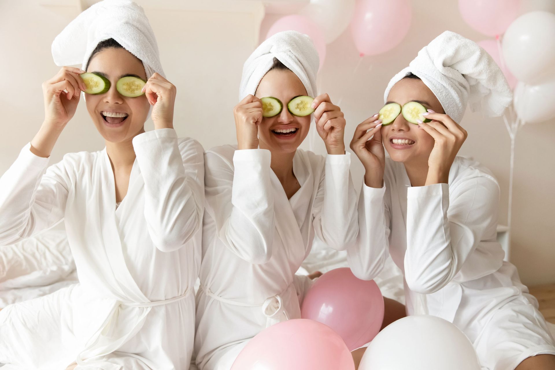 Three women with towels wrapped around their heads are holding cucumber slices in front of their eyes.