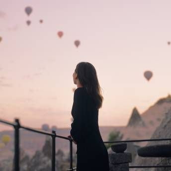 A woman is standing on a balcony looking at hot air balloons flying in the sky.