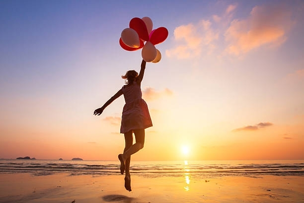A woman is jumping in the air while holding balloons on the beach at sunset.