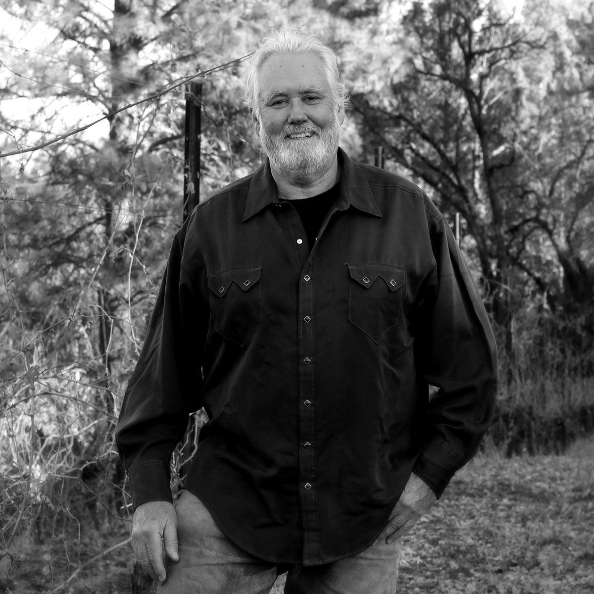 A man with a beard is standing in a field with his hands in his pockets.
