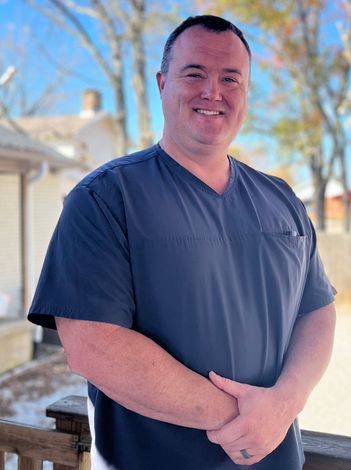 A man in a scrub top is standing on a balcony with his hands folded.