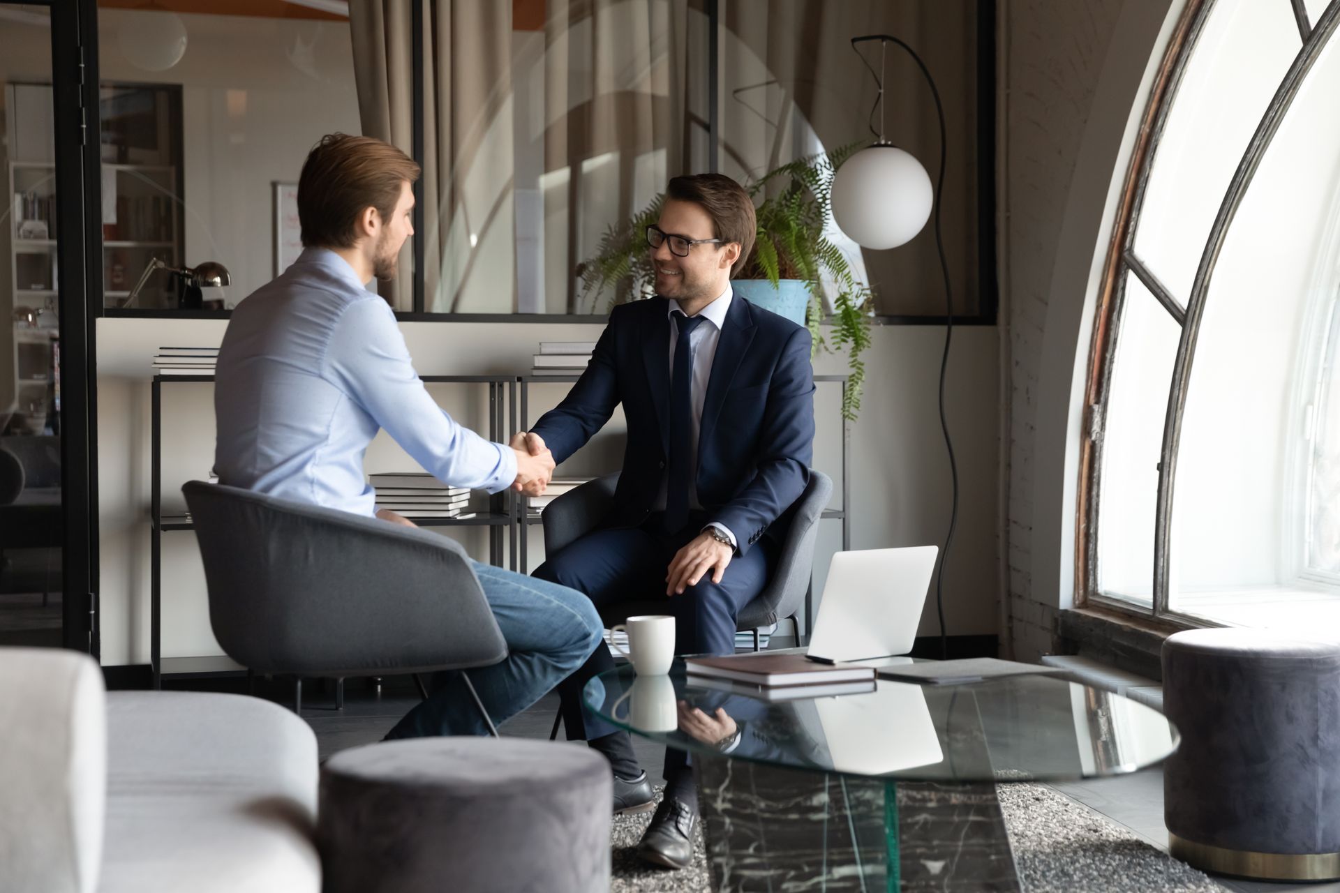 Two men are shaking hands while sitting at a table in a living room.