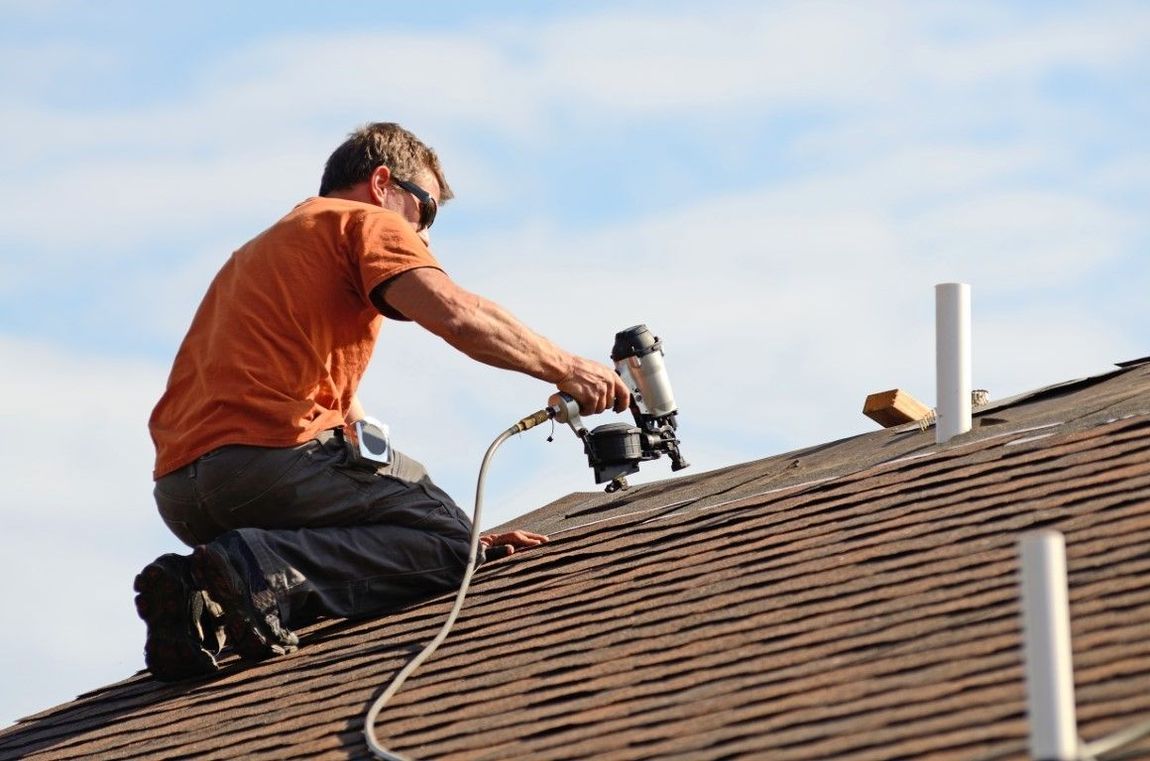 Roofer in orange shirt kneels on a roof, using a nail gun. Blue sky in the background