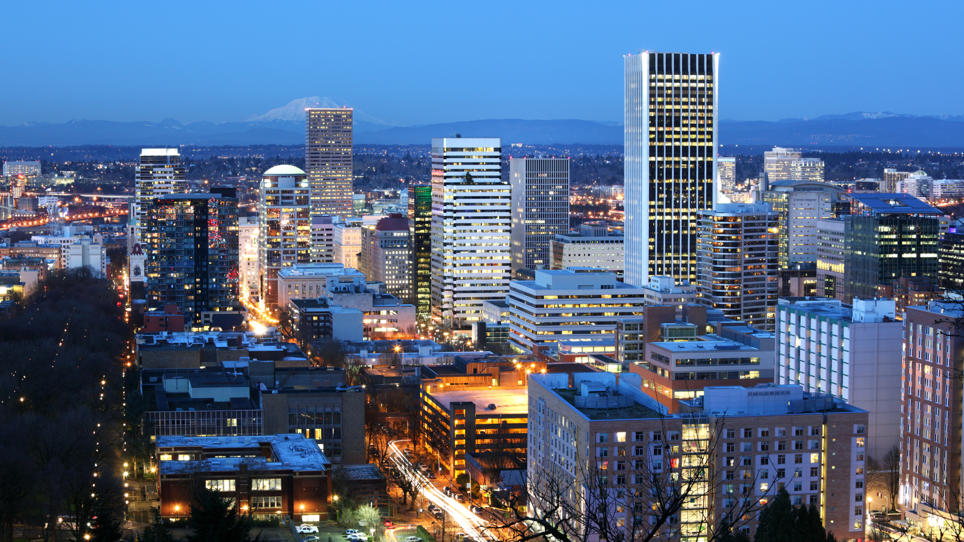 Downtown Portland cityscape at dusk with lit buildings and Mt. Hood in the background.