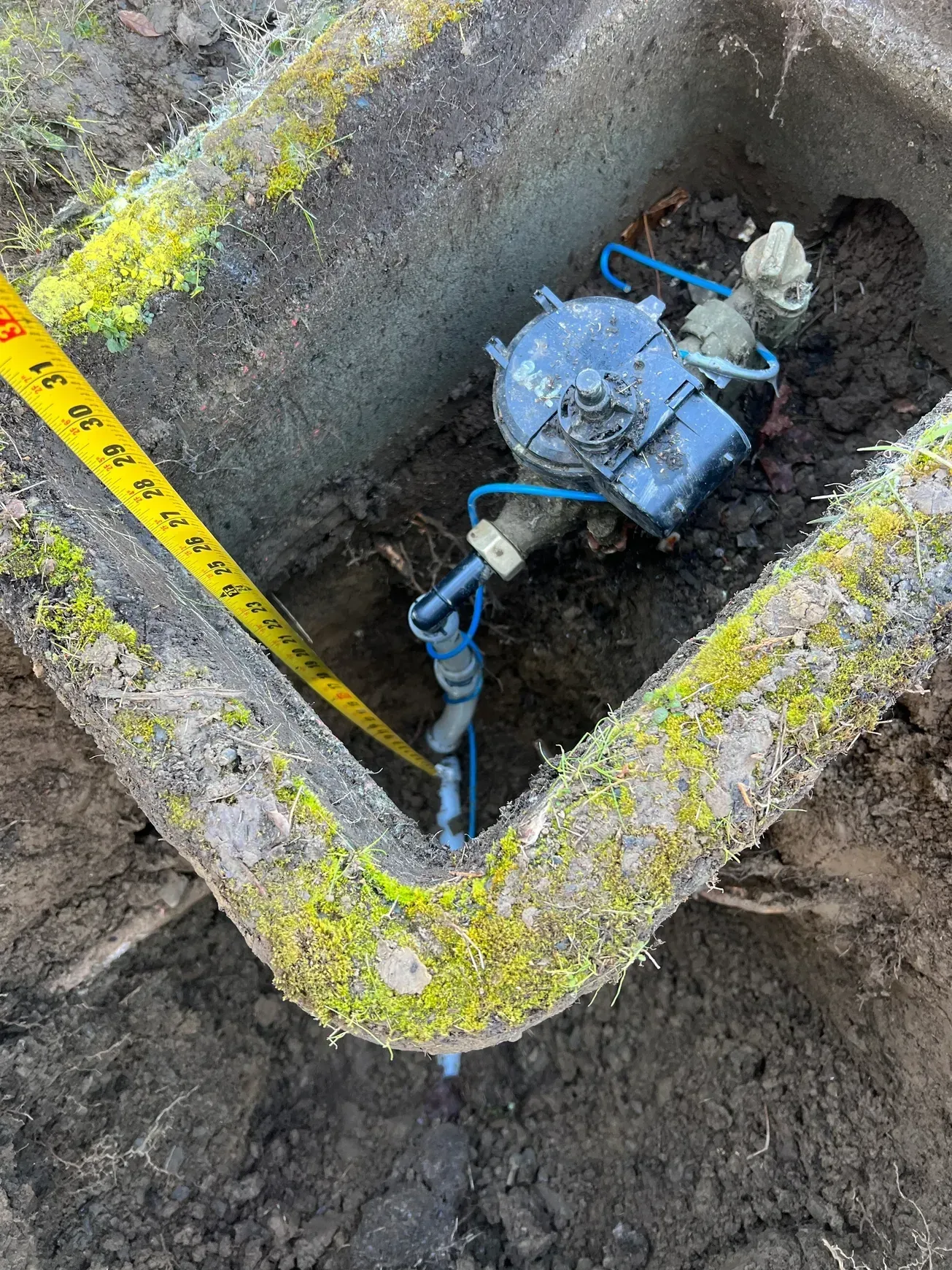 A water meter is sitting in the dirt next to a tape measure.