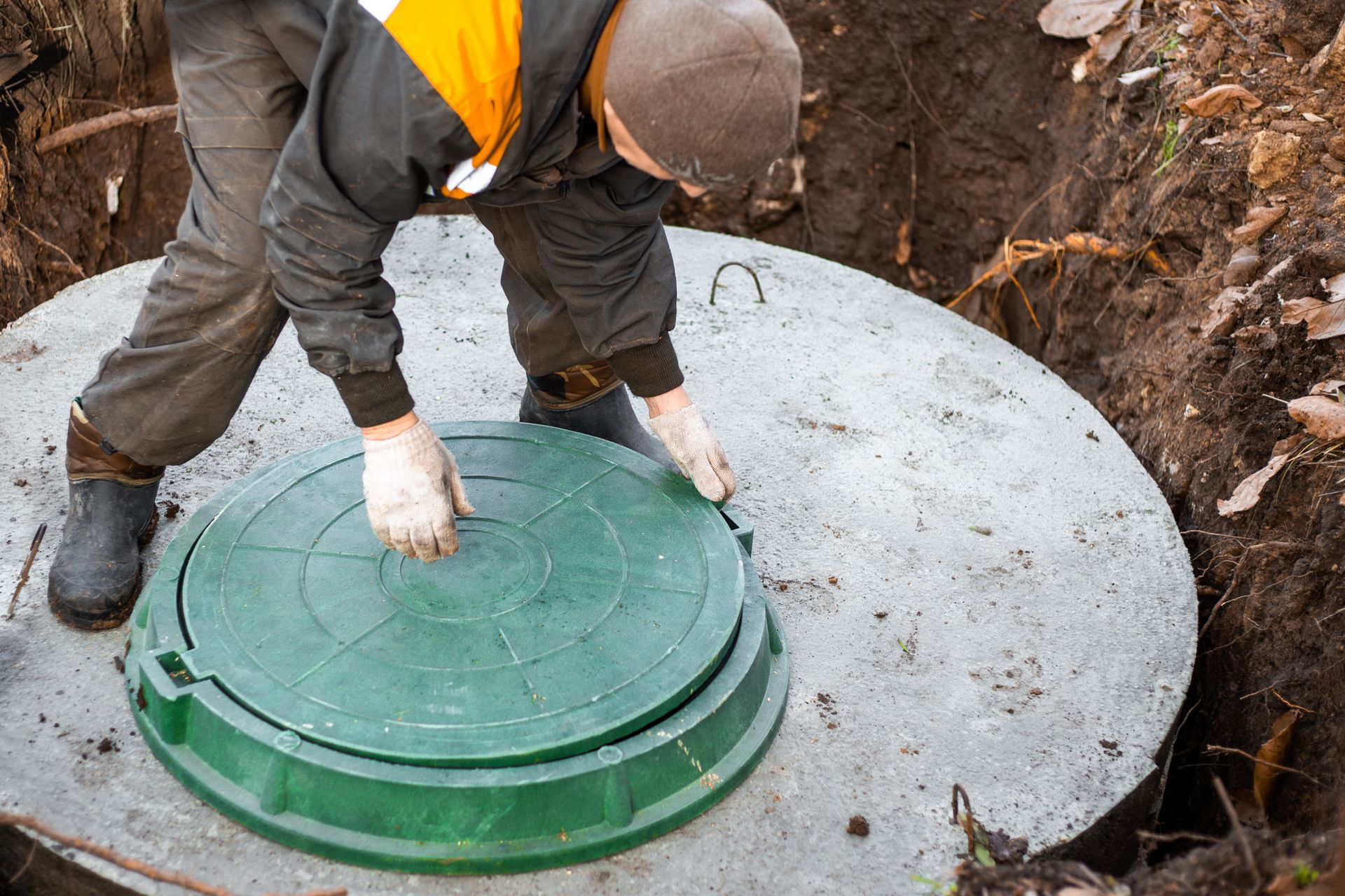 Worker in safety gear opening green septic tank lid outdoors.