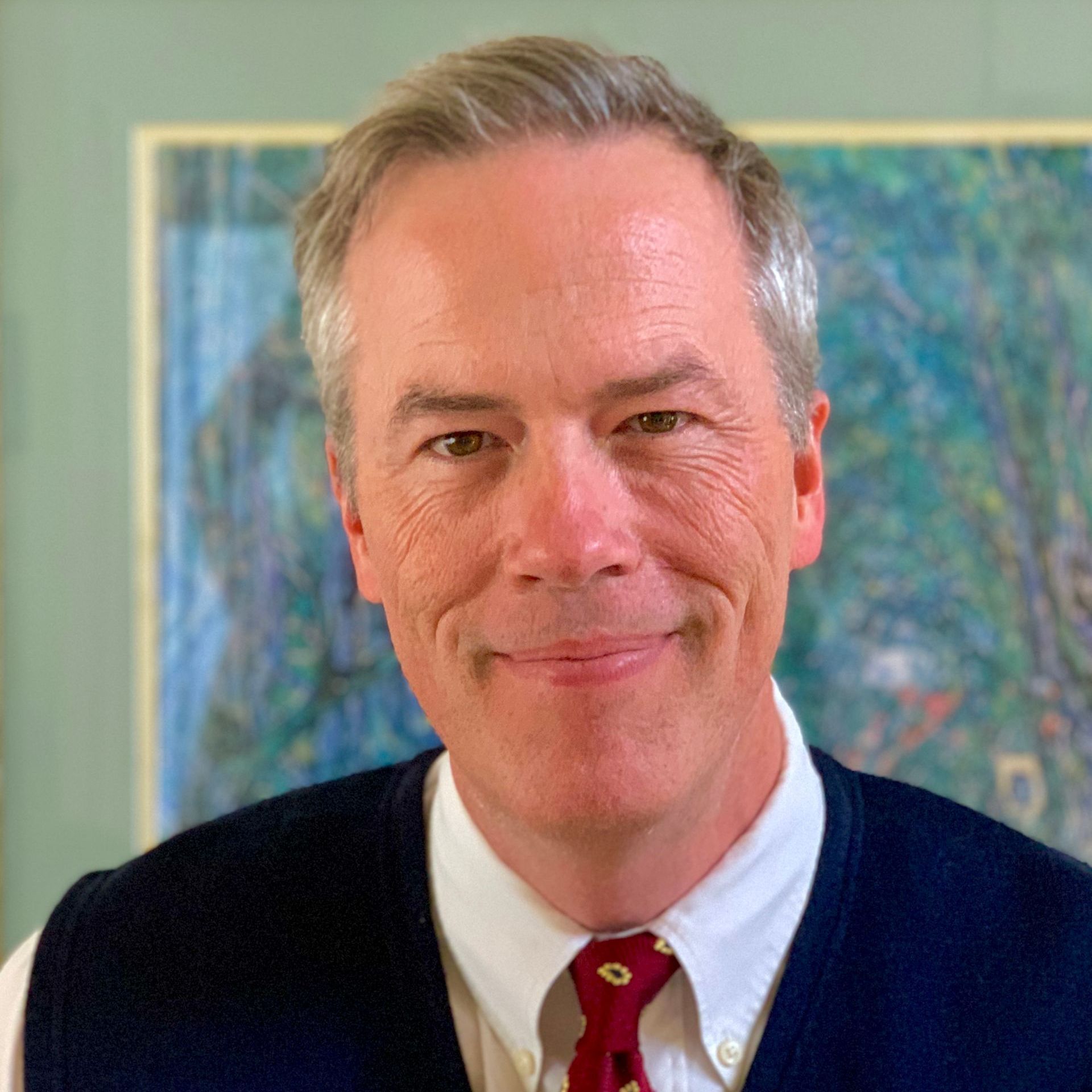 A man wearing a vest and tie is smiling in front of a painting.