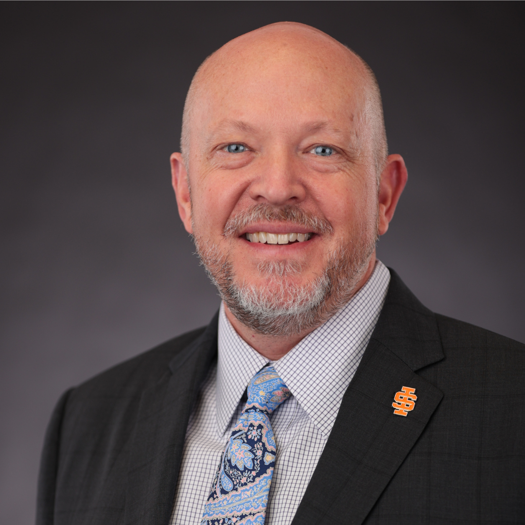 A bald man with a beard wearing a suit and tie is smiling for the camera.