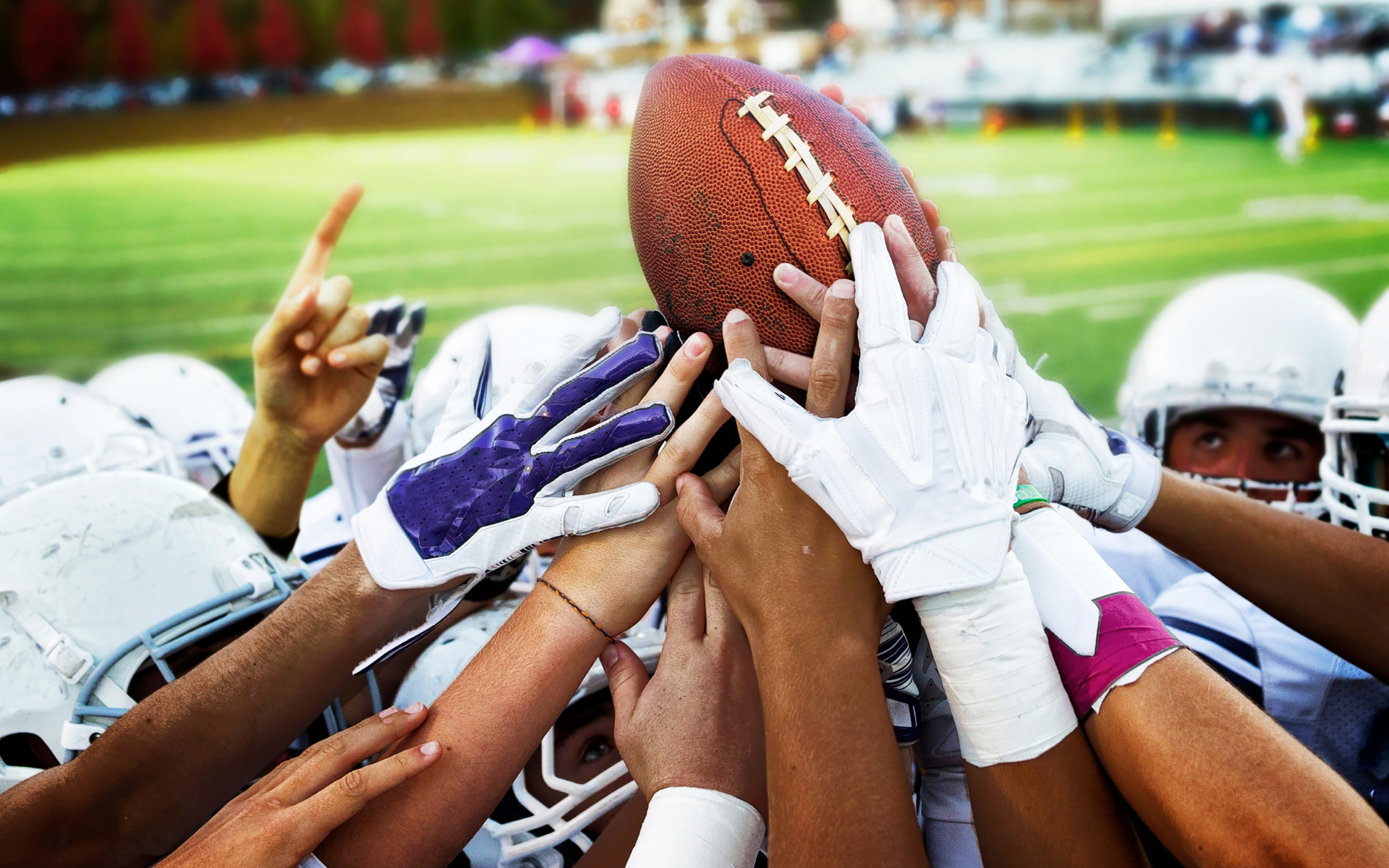 Football team huddle, hands reaching for ball, helmets, and field in background.