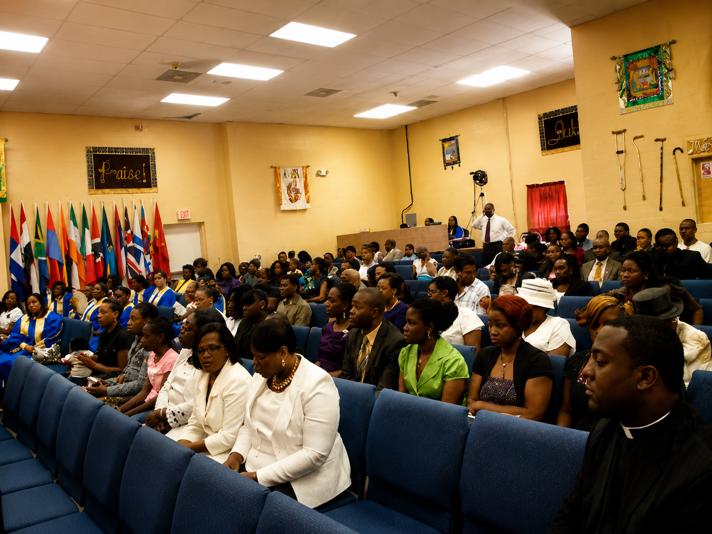 Audience seated in a church or community hall, with rows of blue chairs and colorful flags along the wall.
