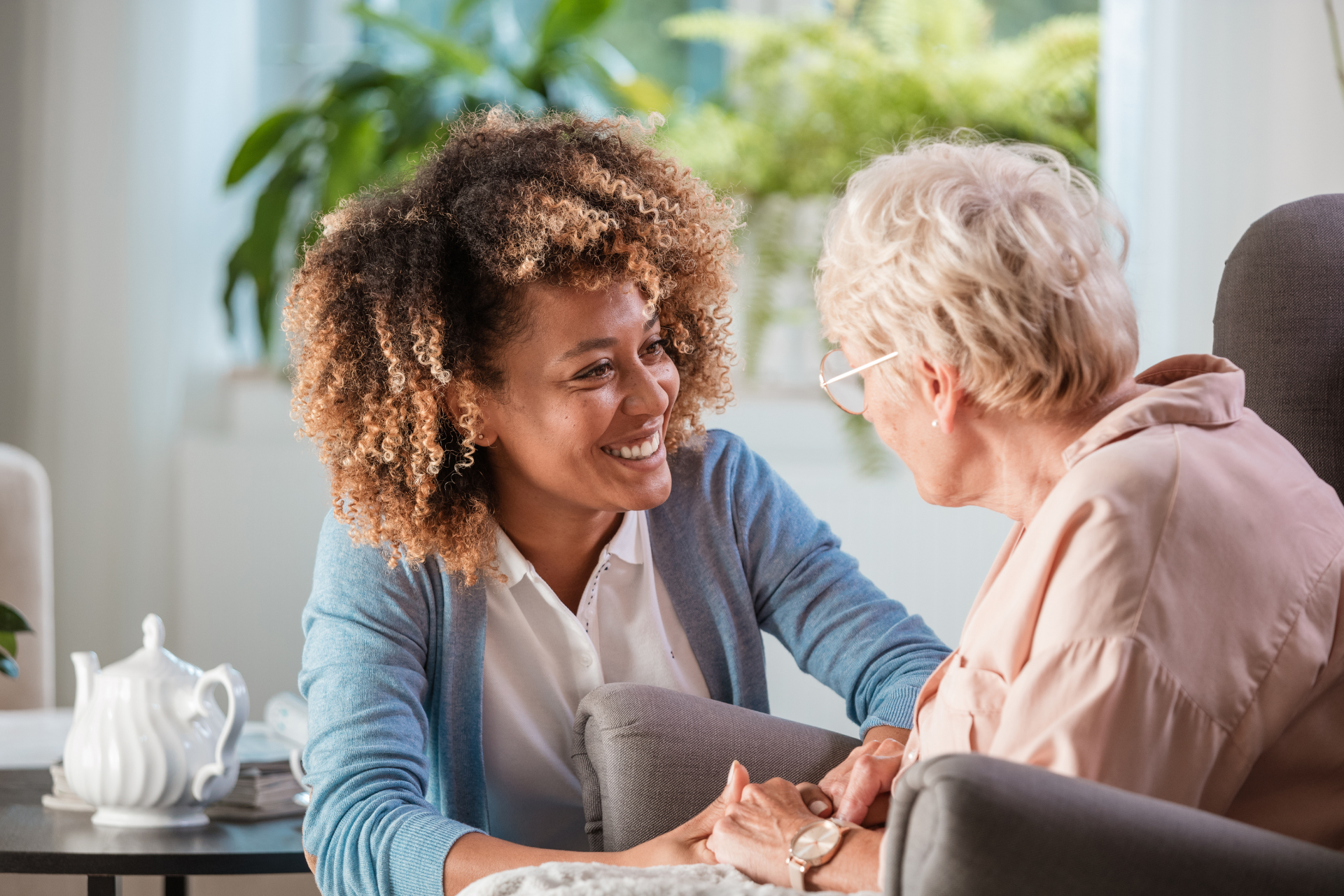 Woman smiles while holding hands with an older person in a living room; teapot on a side table.
