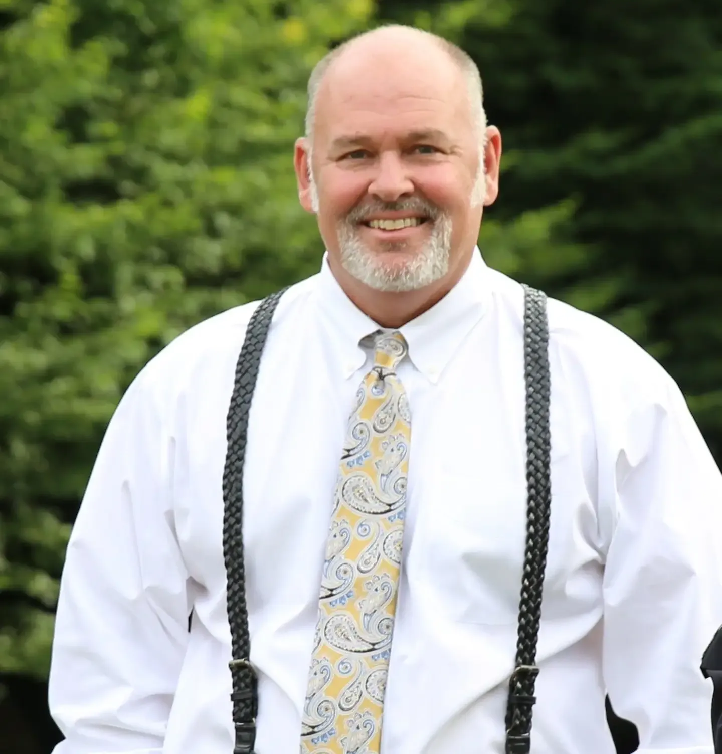Man with a graying beard smiles, wearing a white shirt, patterned tie, and suspenders.