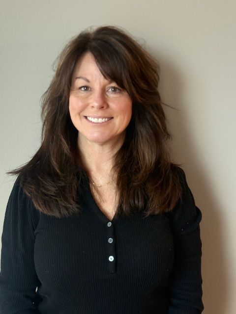 Woman with shoulder-length dark brown hair smiles, wearing a black shirt, against a beige wall.