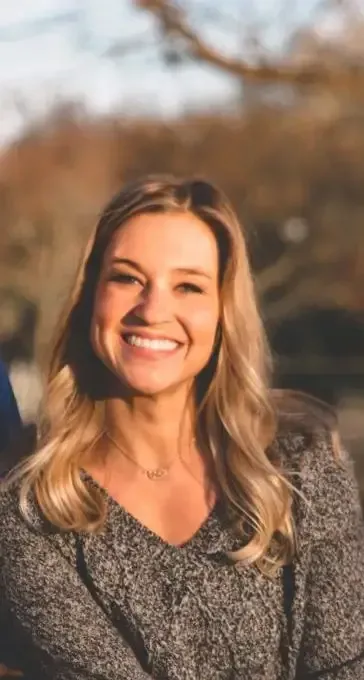 Woman smiling outdoors, wearing a gray sweater. Blonde hair, trees in the background.
