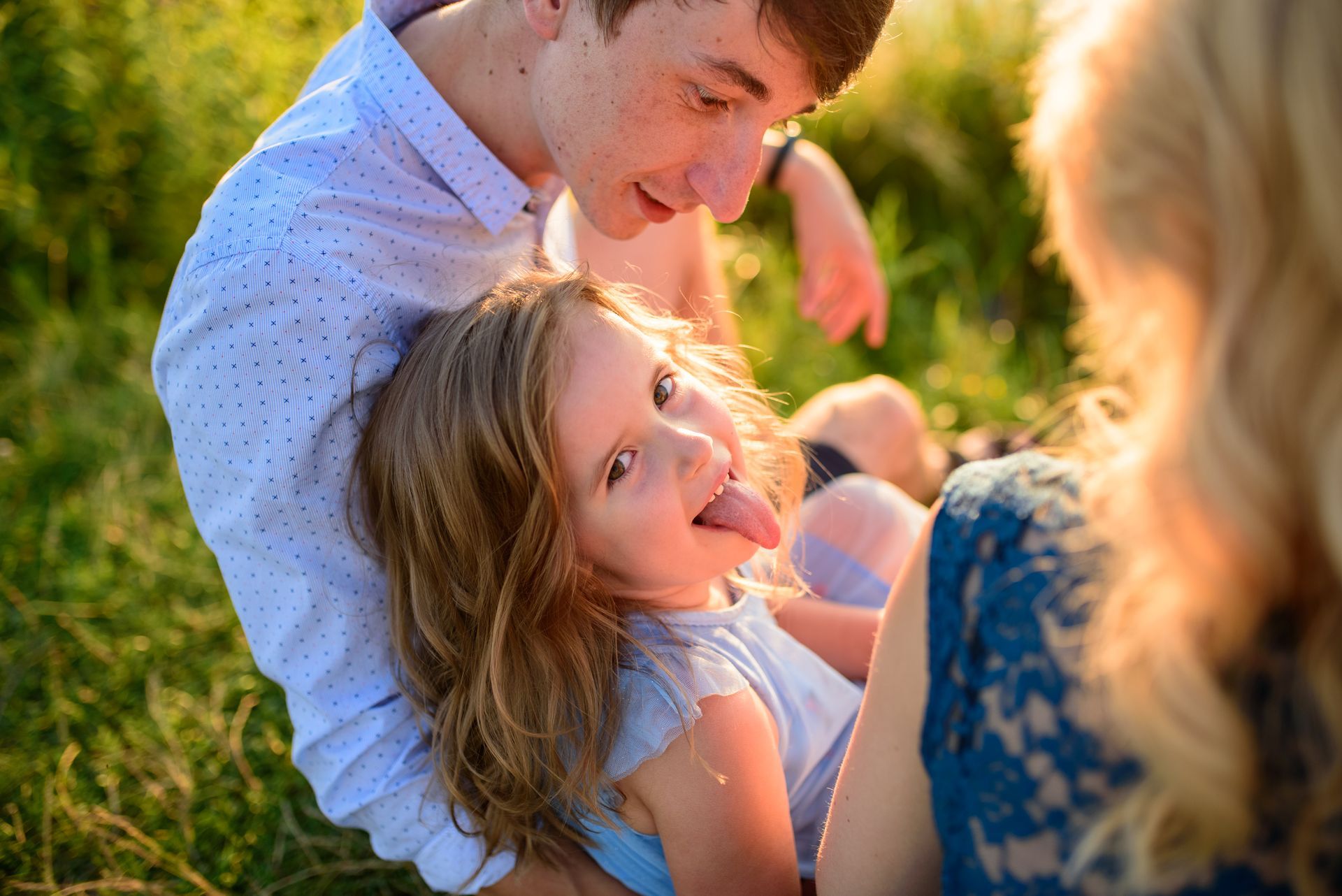 Man holds child, who sticks out her tongue, while seated near a woman outdoors.