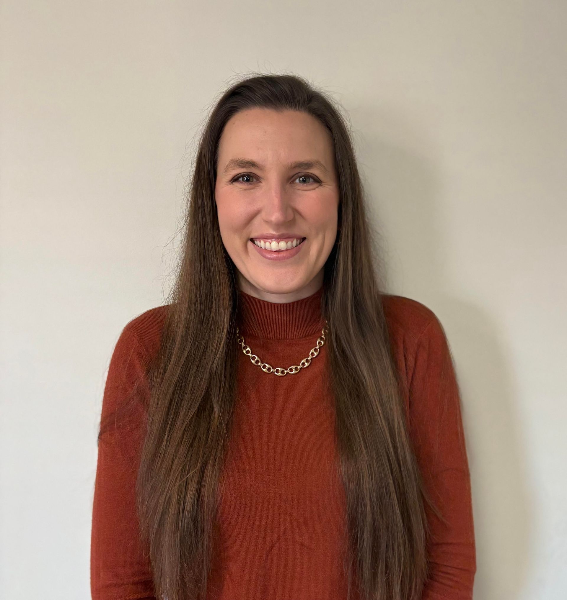 Woman with long brown hair, smiling, wearing an orange turtleneck and gold chain necklace against a light wall.