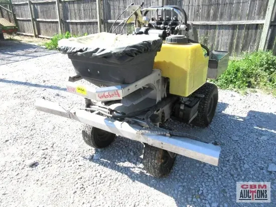 A lawn mower is parked in a gravel lot next to a wooden fence.