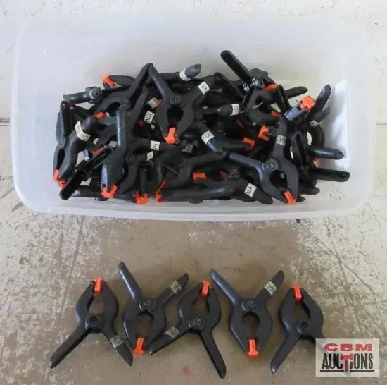 Black spring clamps in a white bin, and six more arranged below on a concrete surface.