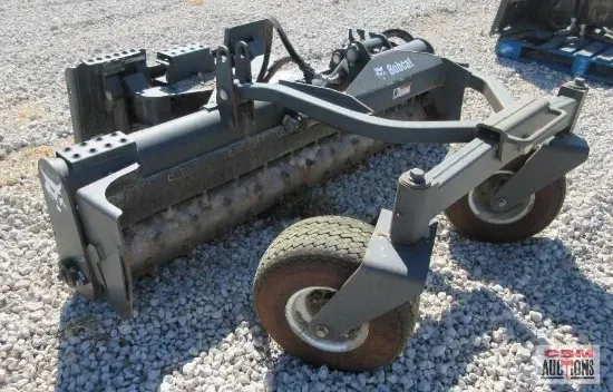 A Bobcat grading attachment on gravel, showing a roller with tires and metal frame.