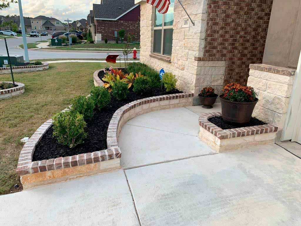 Curved brick-edged flower bed with green plants, black mulch, and a concrete pathway in front of a house.