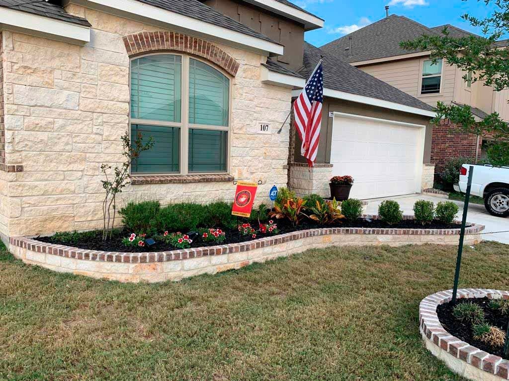 A tan brick house with a flower bed, American flag, and garage.