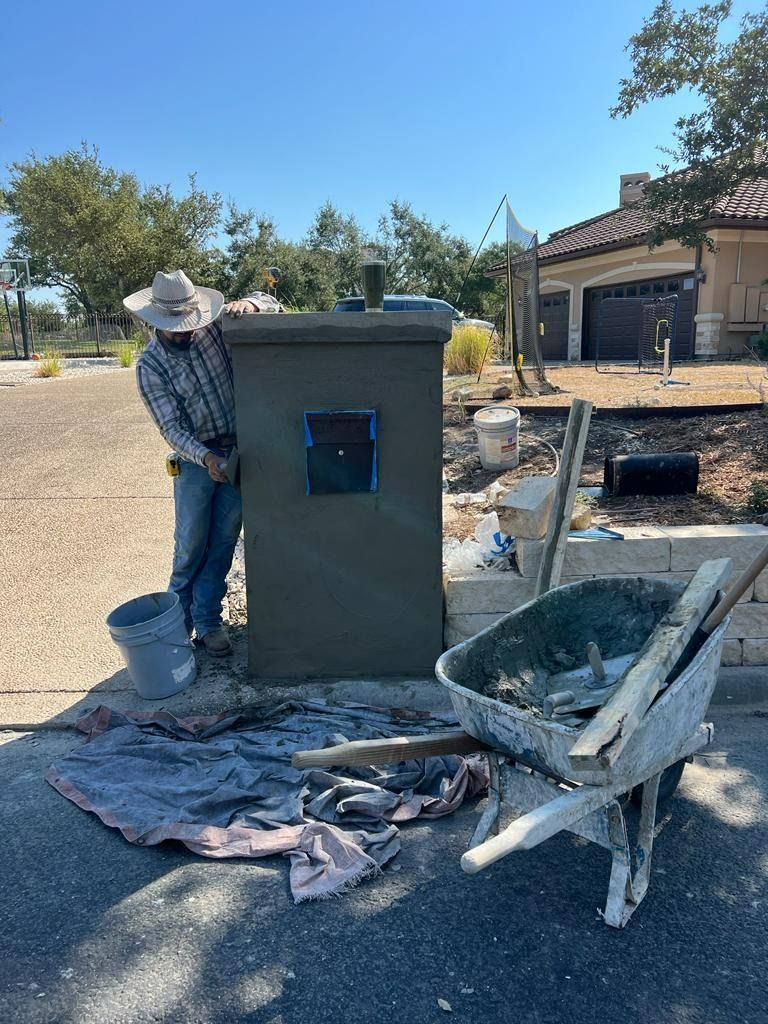 Man applying stucco to a mailbox. Blue sky, wheelbarrow, and bucket present.