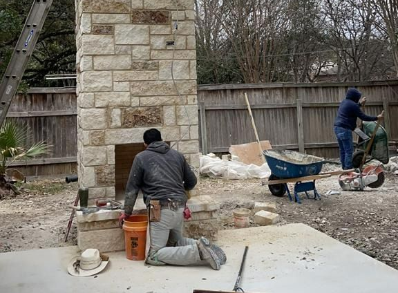 Workers building a stone fireplace outdoors; one kneels, one moves a wheelbarrow, surrounded by tools and materials.