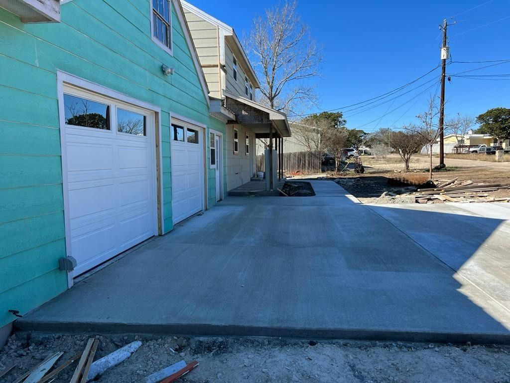 Concrete driveway leads to a two-story building with a garage and teal siding. Sunny day.