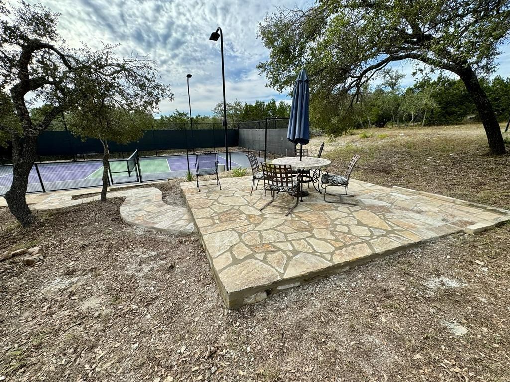 Stone patio with table and chairs, overlooking a tennis court and trees.