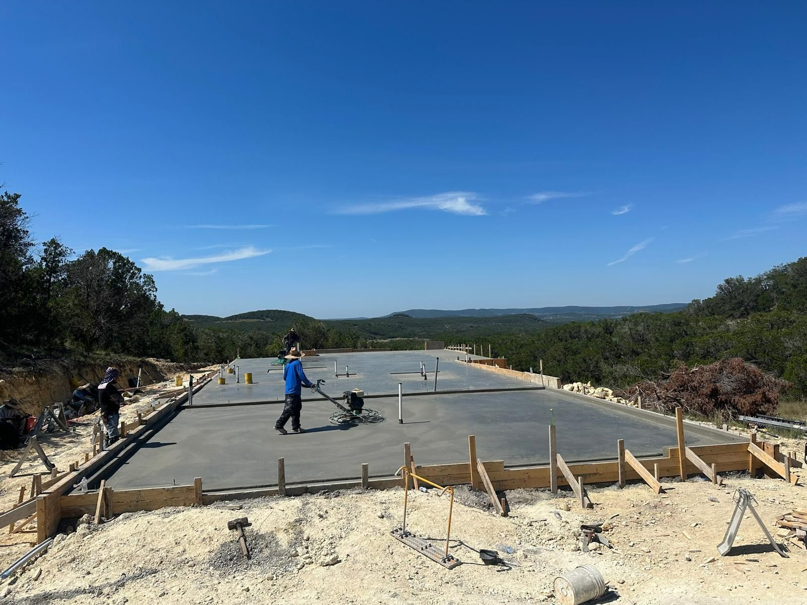 Workers leveling fresh concrete on a sunny day. Wooden forms outline the large, flat surface with a scenic background.