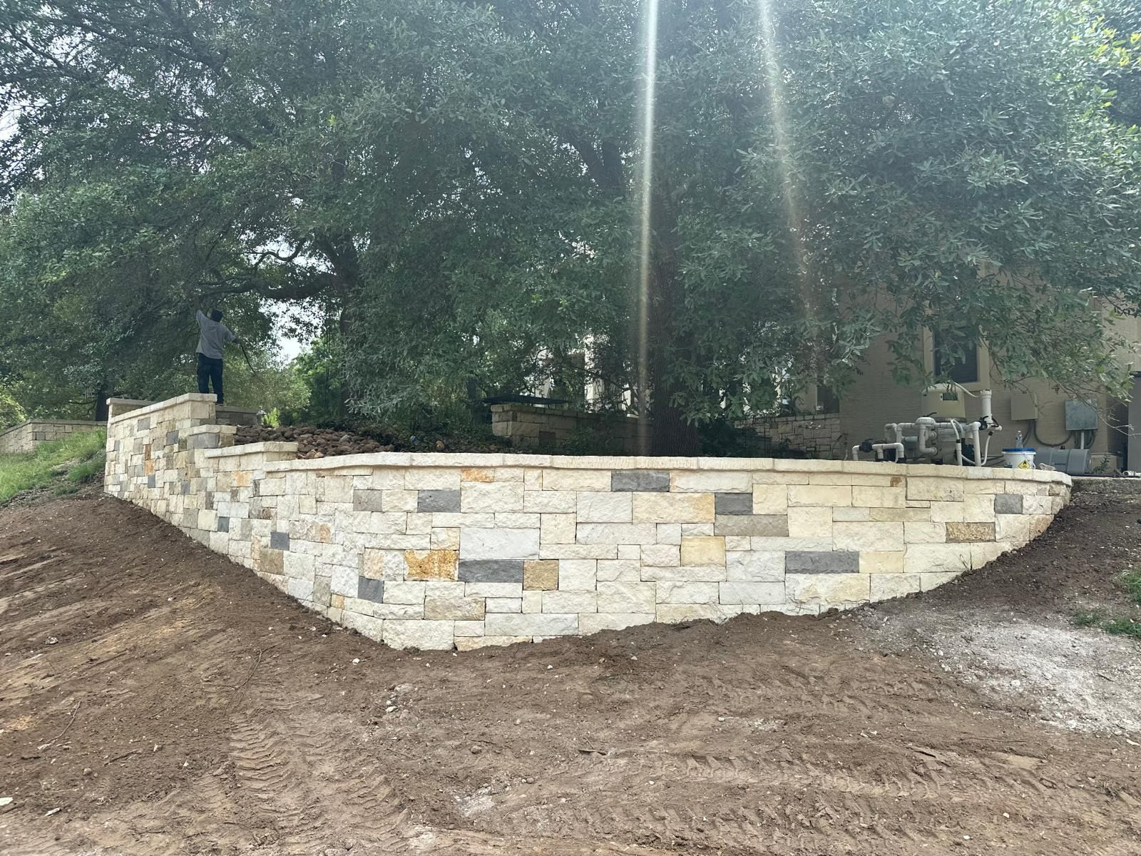 Stone retaining wall in front of a tree, built on a slope with person standing on top.