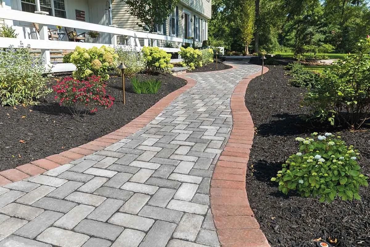 Brick pathway with dark mulch borders, leading to a white house, surrounded by landscaping.