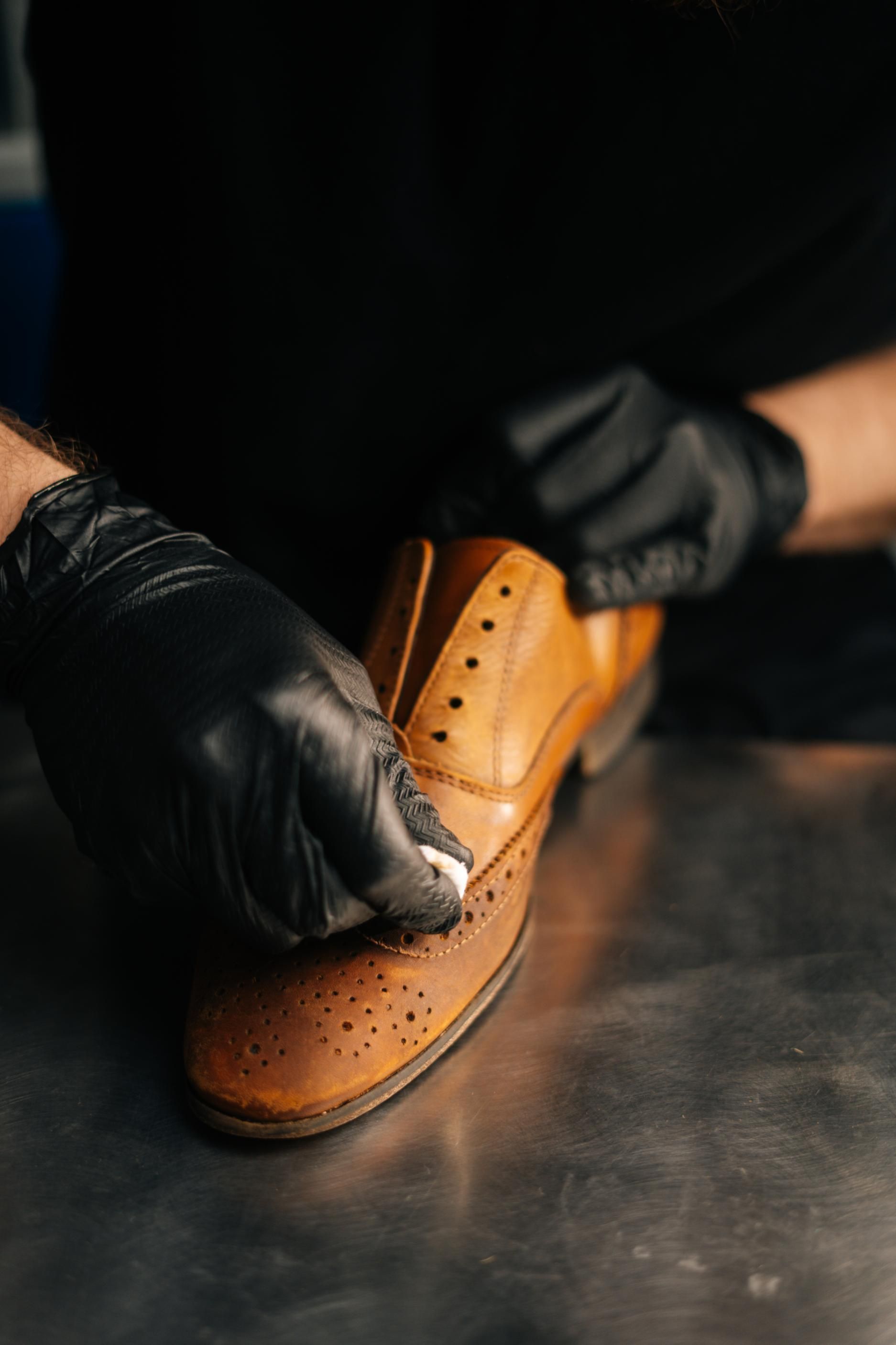 A person wearing black gloves is cleaning a pair of brown shoes.