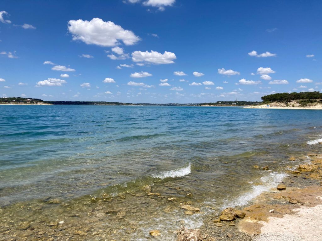 Calm blue ocean shoreline with clear water, rocky beach, and a bright sky with scattered clouds.