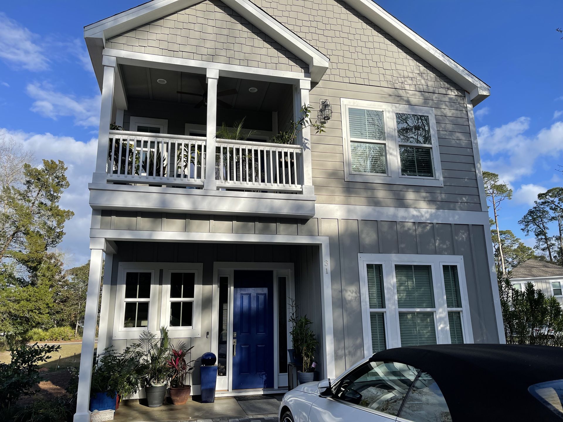 Two-story gray house with a blue front door and balcony. White car parked in the foreground.