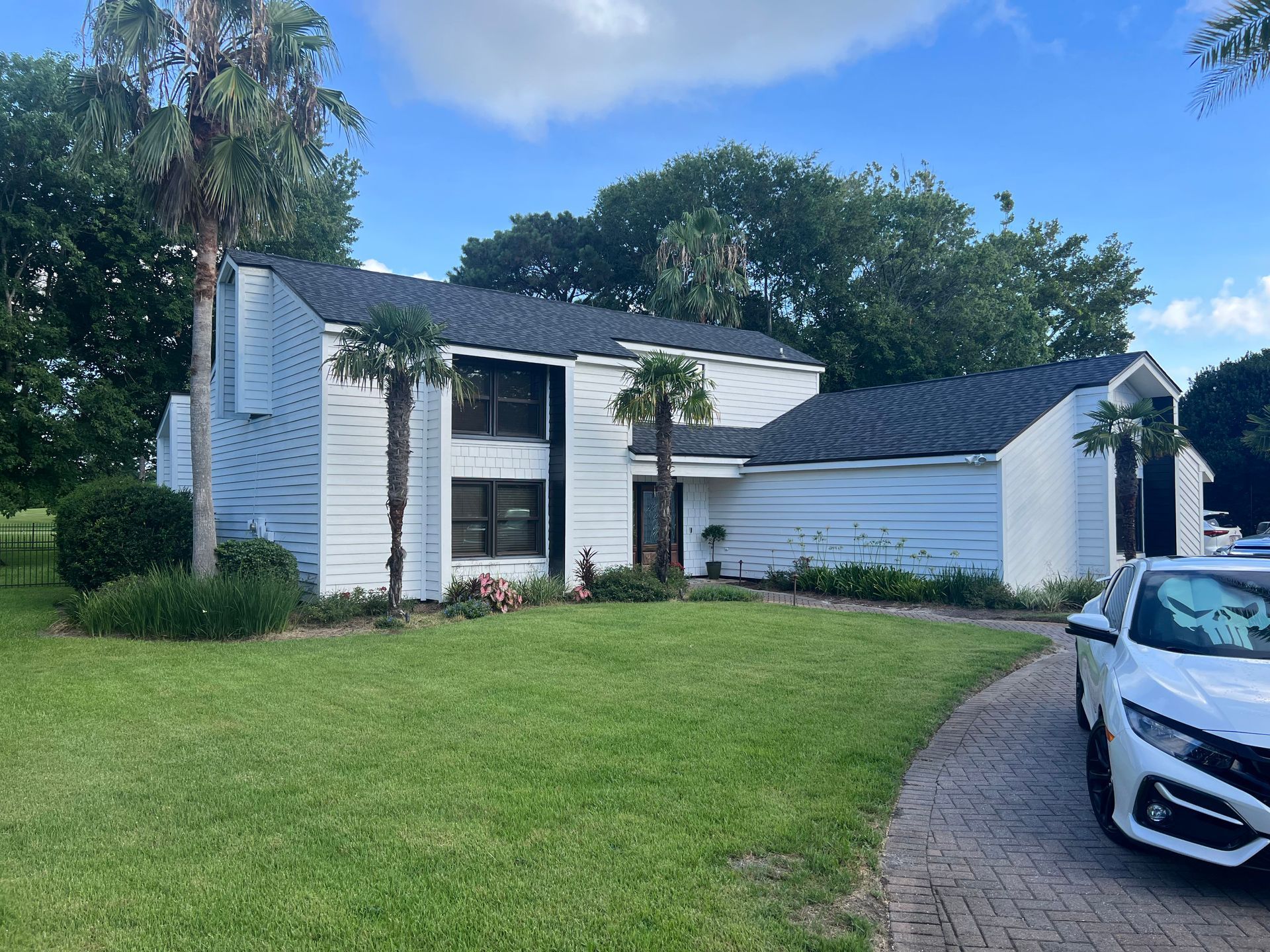 White two-story house with black roof, palm trees, and a parked car on a grassy lawn.
