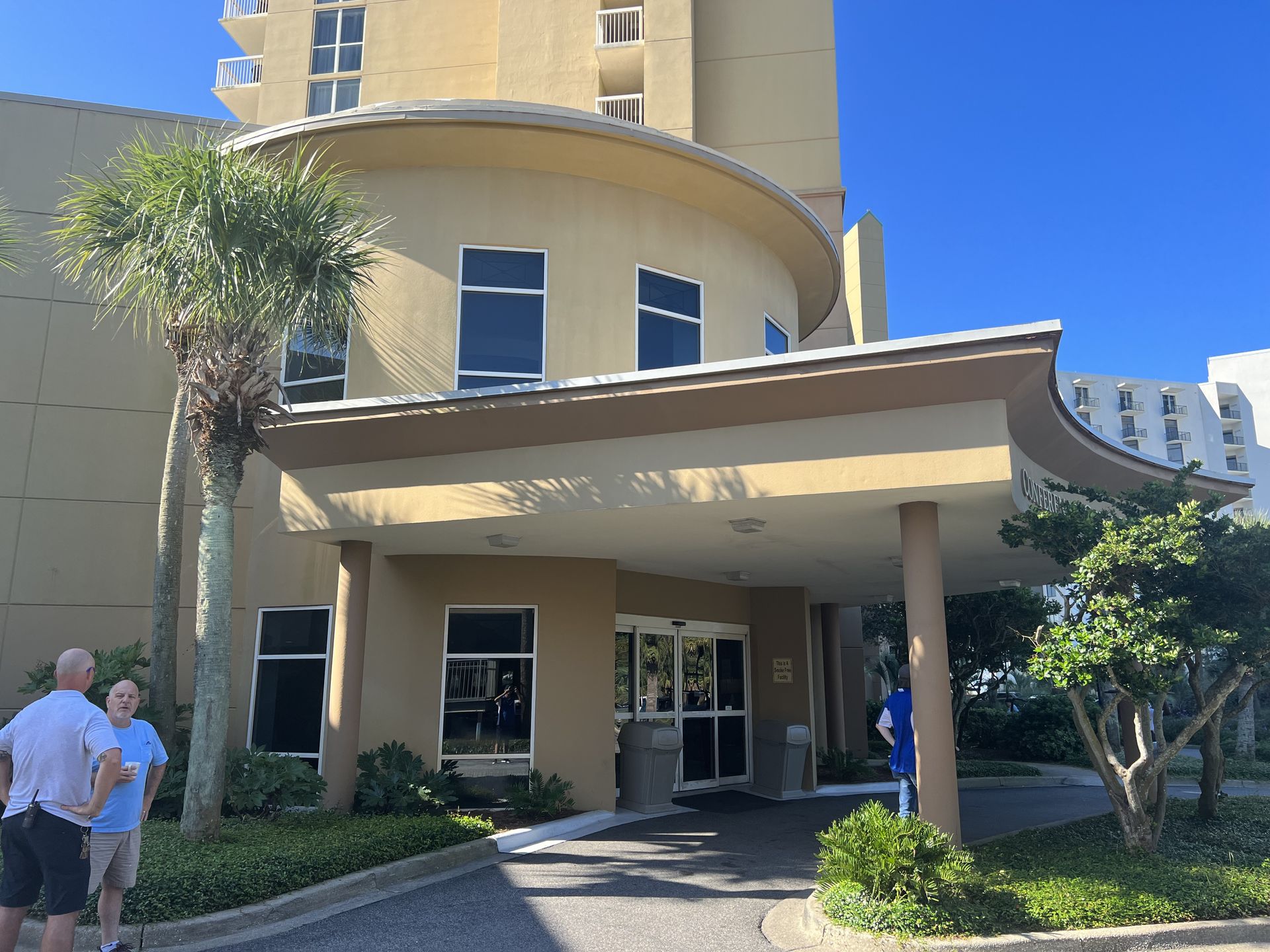 Entrance to a light tan building with a curved roof and glass doors, palm tree, two people standing.