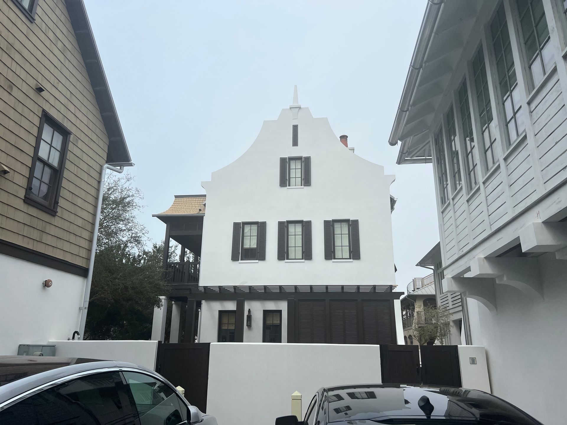 White stucco building with a distinctive peaked roof, flanked by wooden houses, under overcast skies.