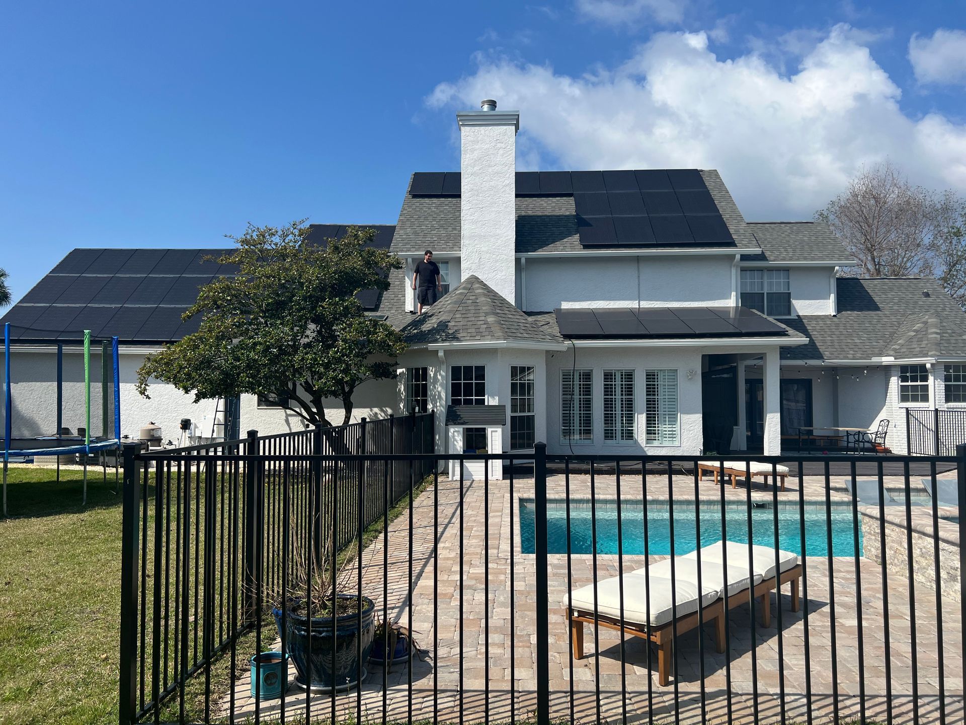 House with solar panels on roof, swimming pool, black fence, and blue sky.