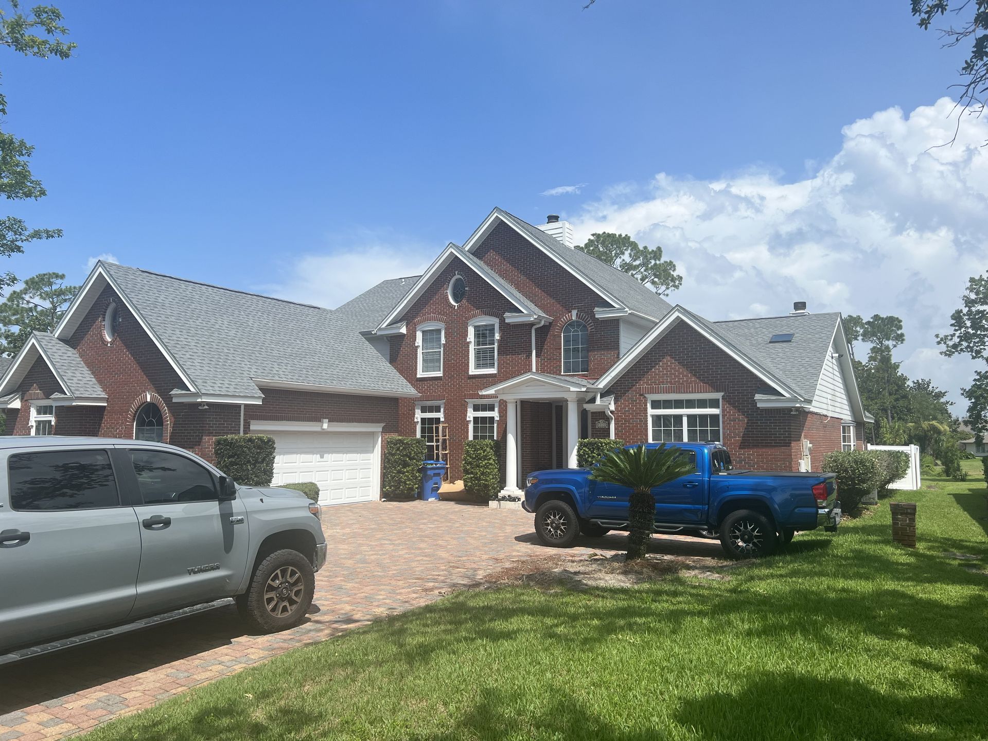 Two-story brick house with gray roof, driveway, two trucks, and green lawn under a blue sky.