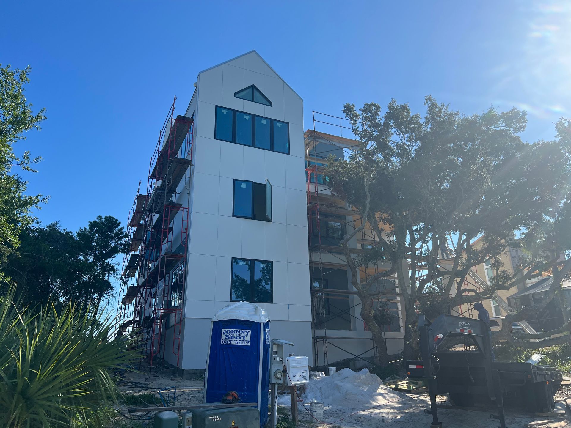 Multi-story building under construction with scaffolding, a blue portable toilet, and trees against a blue sky.