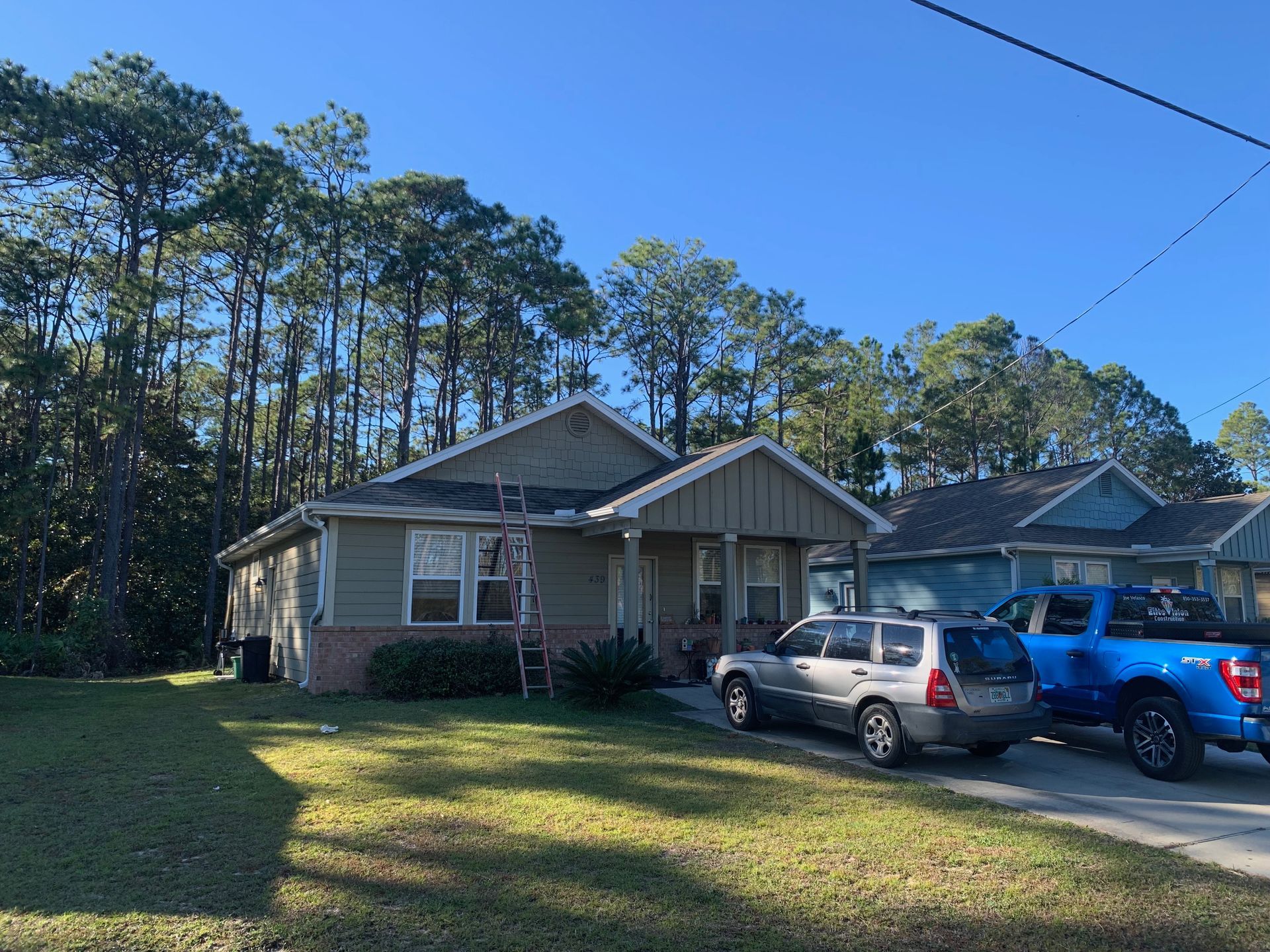 Green house with white trim, ladder on roof. Cars parked in driveway under blue sky.
