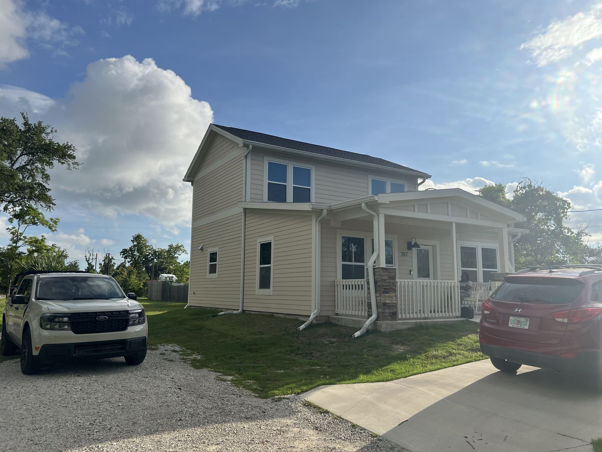 Two-story house with beige siding, porch, and two vehicles parked in the driveway under a partly cloudy sky.