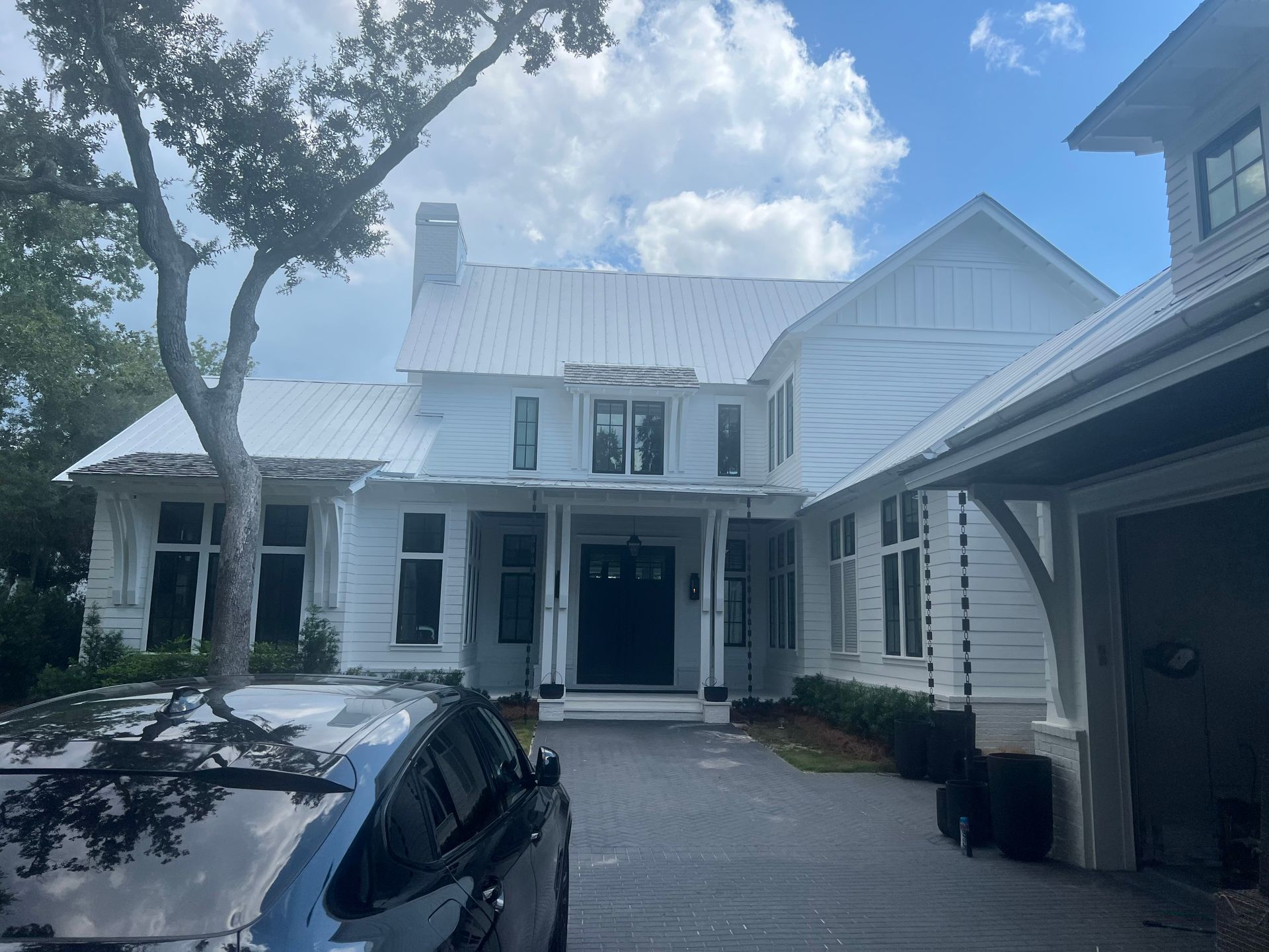 White house with metal roof; dark driveway, black car in foreground, blue sky.