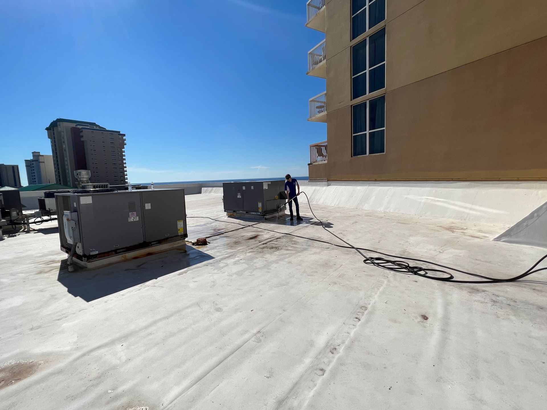 Person cleaning HVAC equipment on a flat rooftop of a beige building on a sunny day.